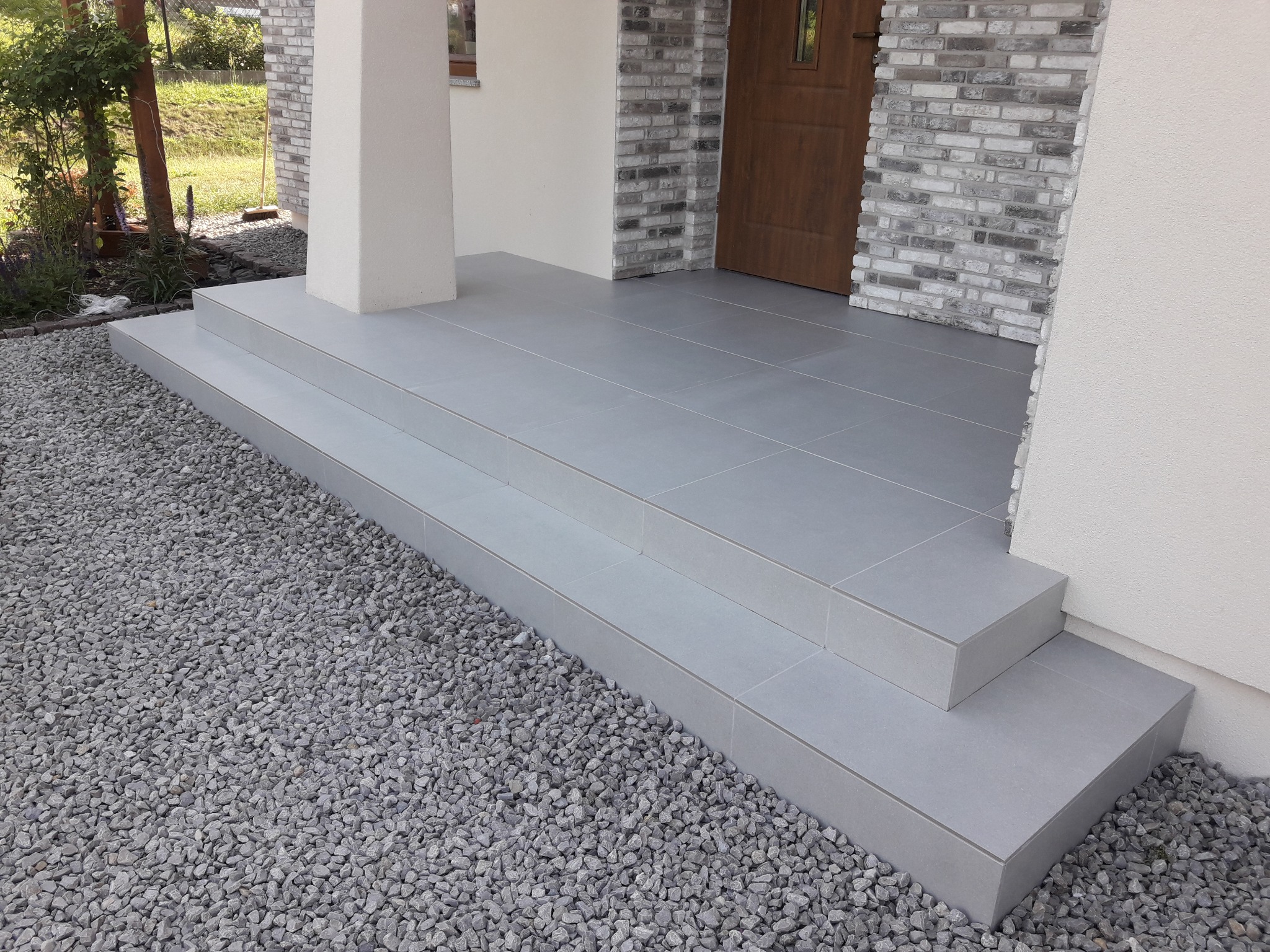 Wykończone schody wejściowe do domu, pokryte szarymi płytkami ceramicznymi, otoczone szarym żwirem, z widoczną częścią elewacji z cegły i drewnianymi drzwiami.