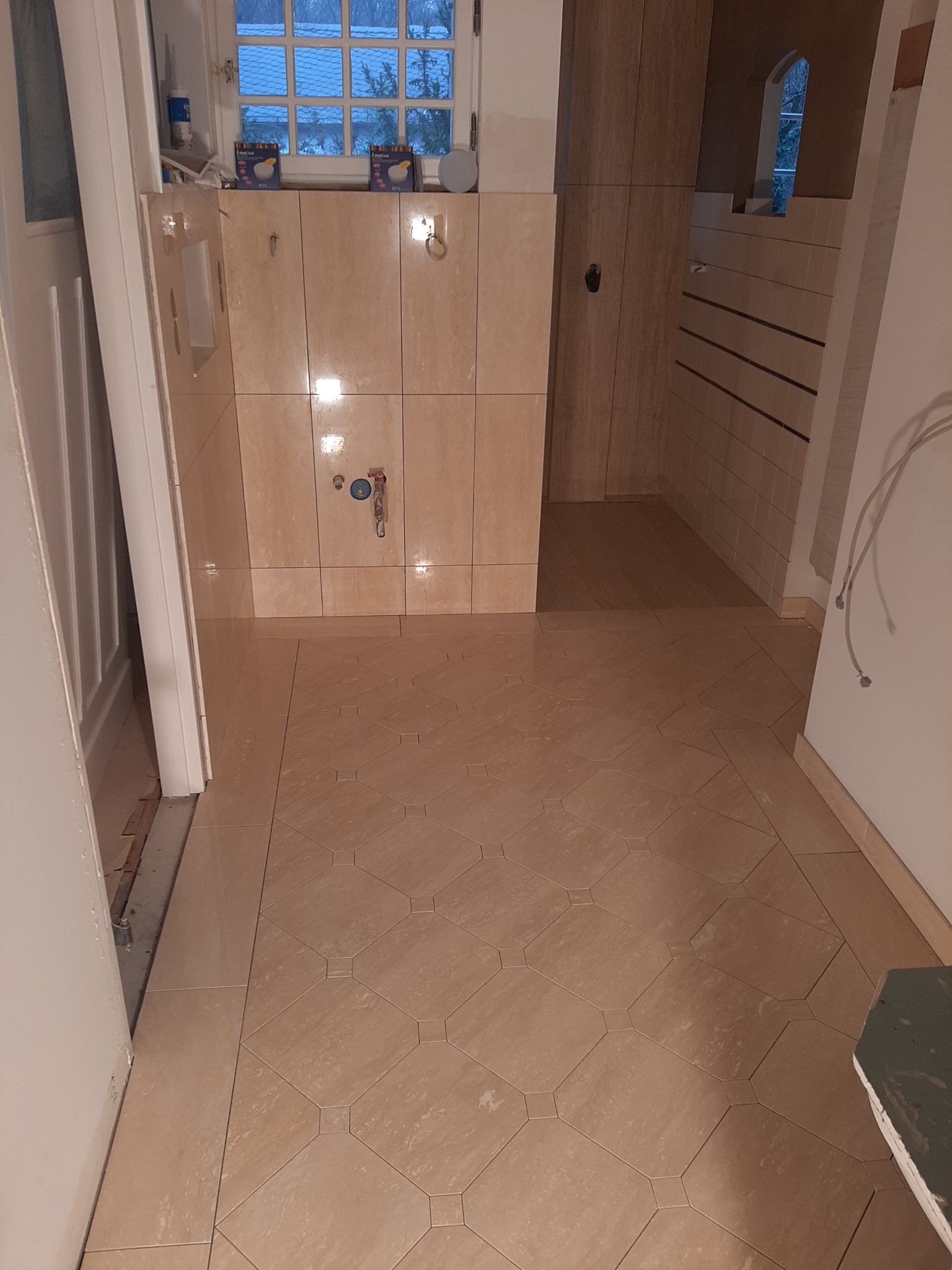 Ukończona łazienka z beżowymi płytkami na ścianach i podłodze w romby, widoczne przyłącza wodne, okno z widokiem na zewnątrz i częściowo otwarte białe drzwi.