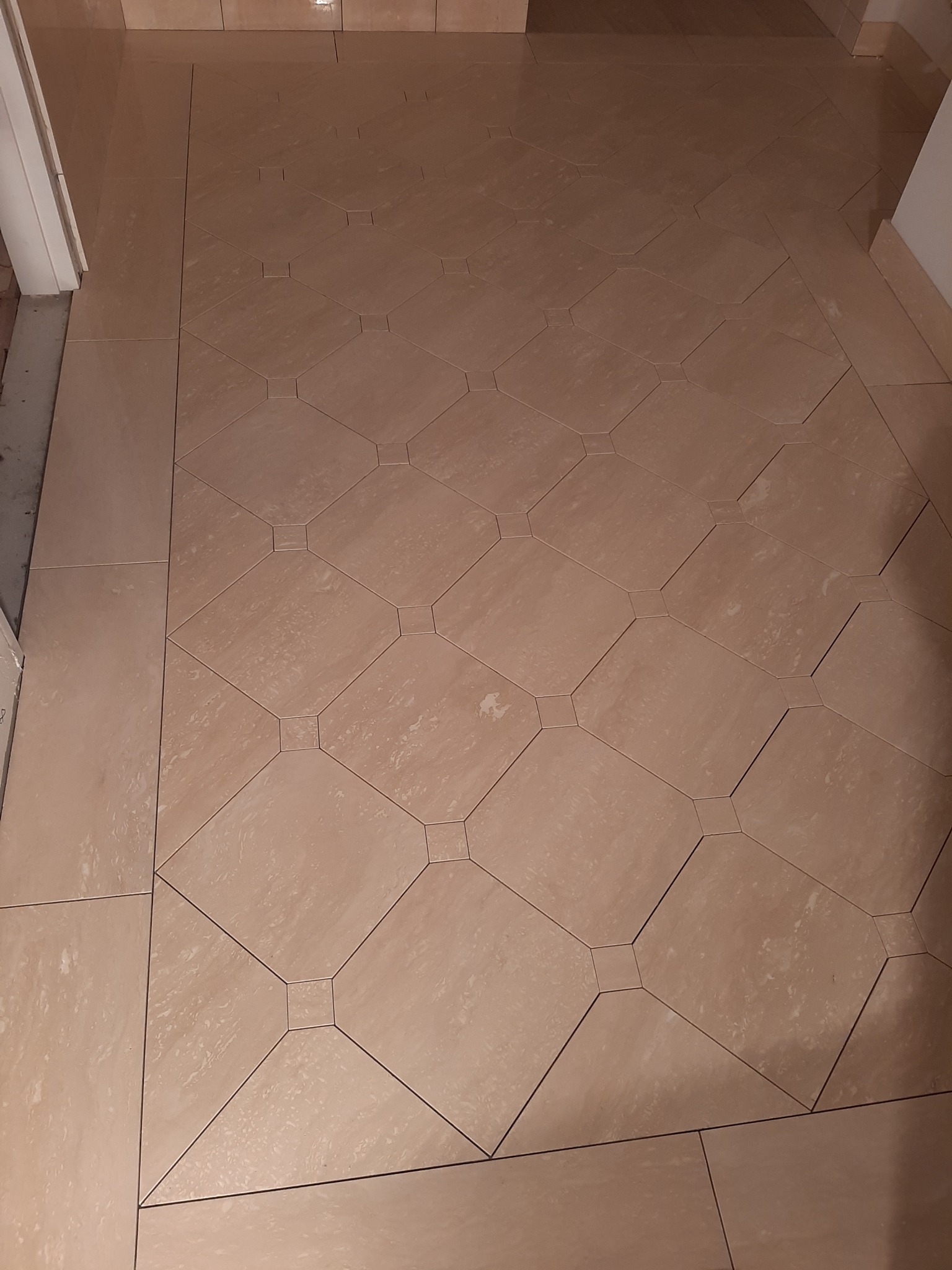Beżowa podłoga wyłożona płytkami w geometryczny wzór, z widocznymi fugami i krawędzią przy ścianie. Widoczne niedoskonałości ułożenia płytek przy progu drzwi.