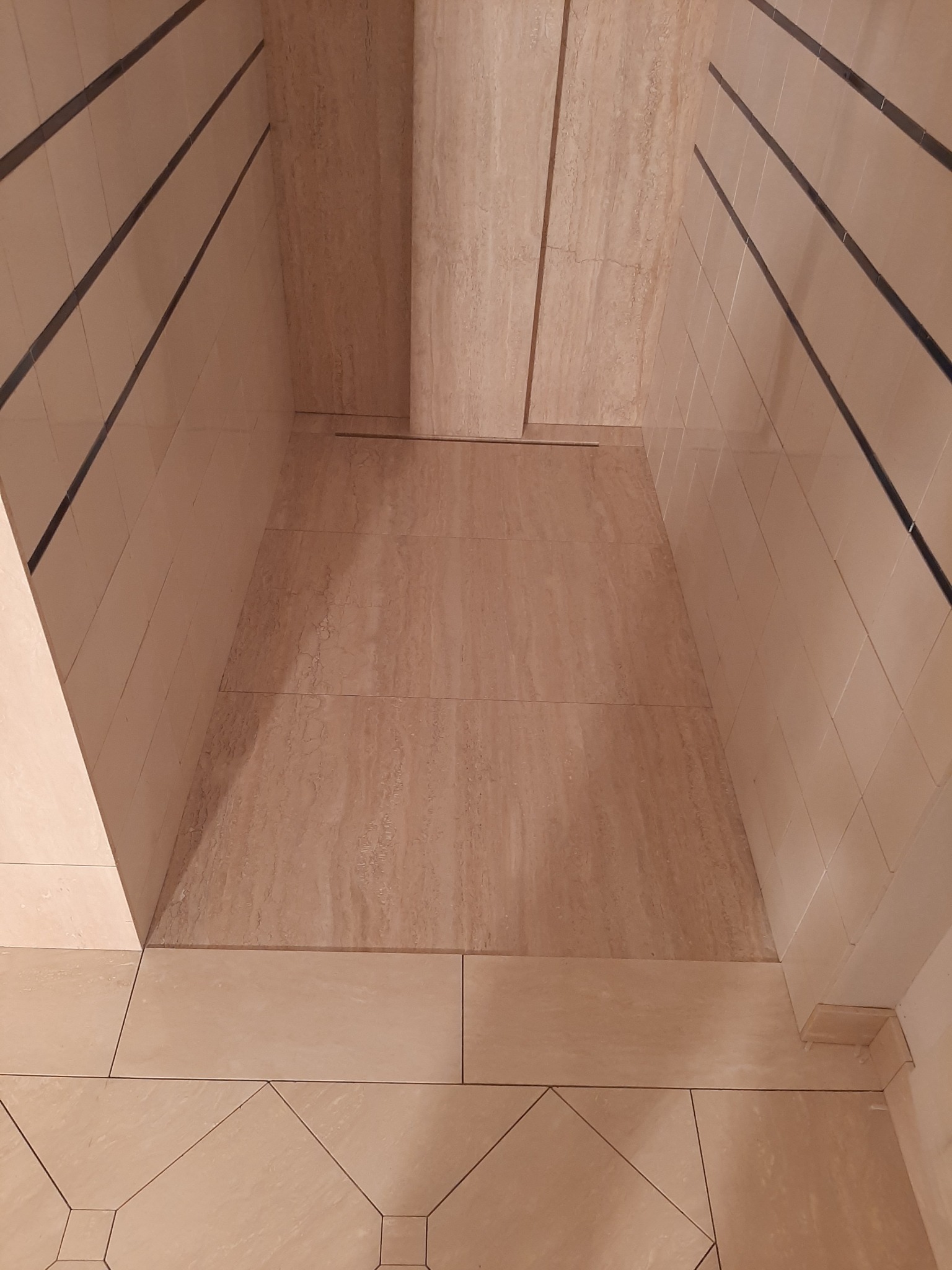 Beżowa kabina prysznicowa wyłożona płytkami imitującymi trawertyn, z czarnymi akcentami na górze płytek ściennych, widok z góry na wykończoną podłogę i ściany.