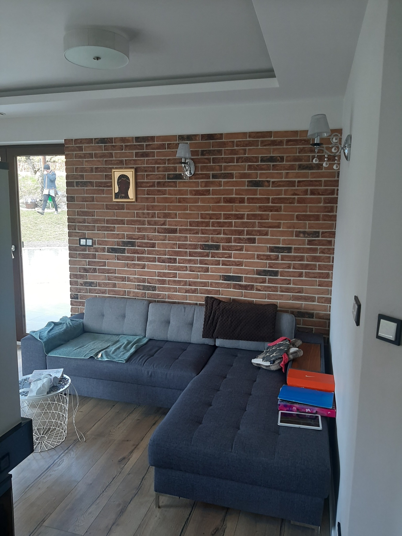 Salon z szarą sofą narożną, ścianą z cegły, drewnianą podłogą i widokiem na ogród przez otwarte drzwi balkonowe.