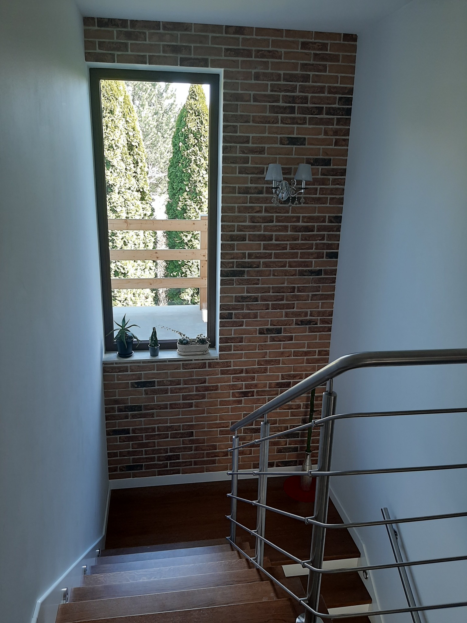 Klatka schodowa z drewnianymi schodami, ceglaną ścianą z oknem i metalową balustradą, oświetlona kinkietem w stylu retro.