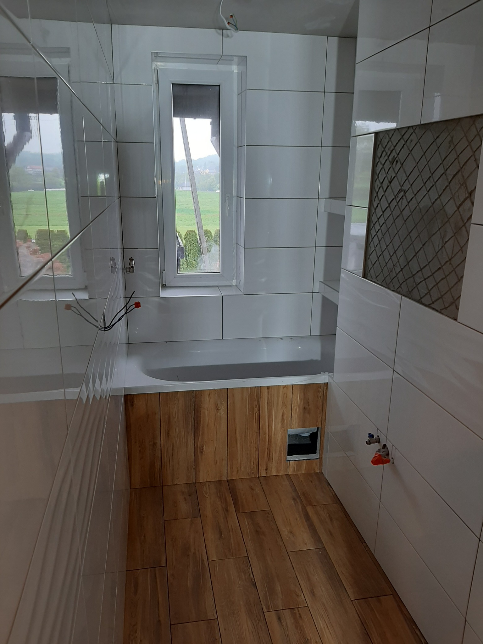 Wnętrze łazienki w trakcie wykańczania: białe płytki ścienne, drewnopodobne panele podłogowe, obudowa wanny z drewnopodobnych paneli, okno z widokiem na zewnątrz, widoczne instalacje wodne...