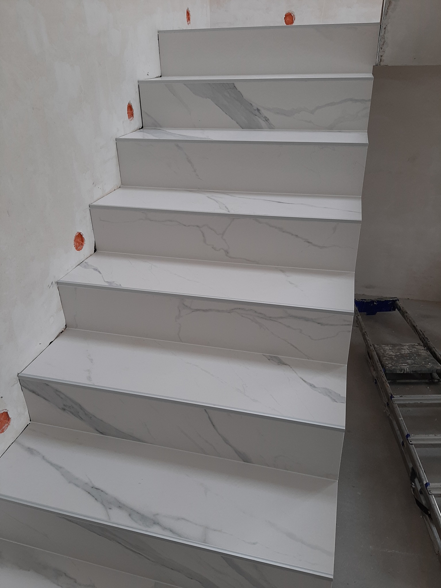 Schody wyłożone białymi płytkami imitującymi marmur, z aluminiowymi listwami krawędziowymi, widoczne niedokończone ściany i drabina w tle.