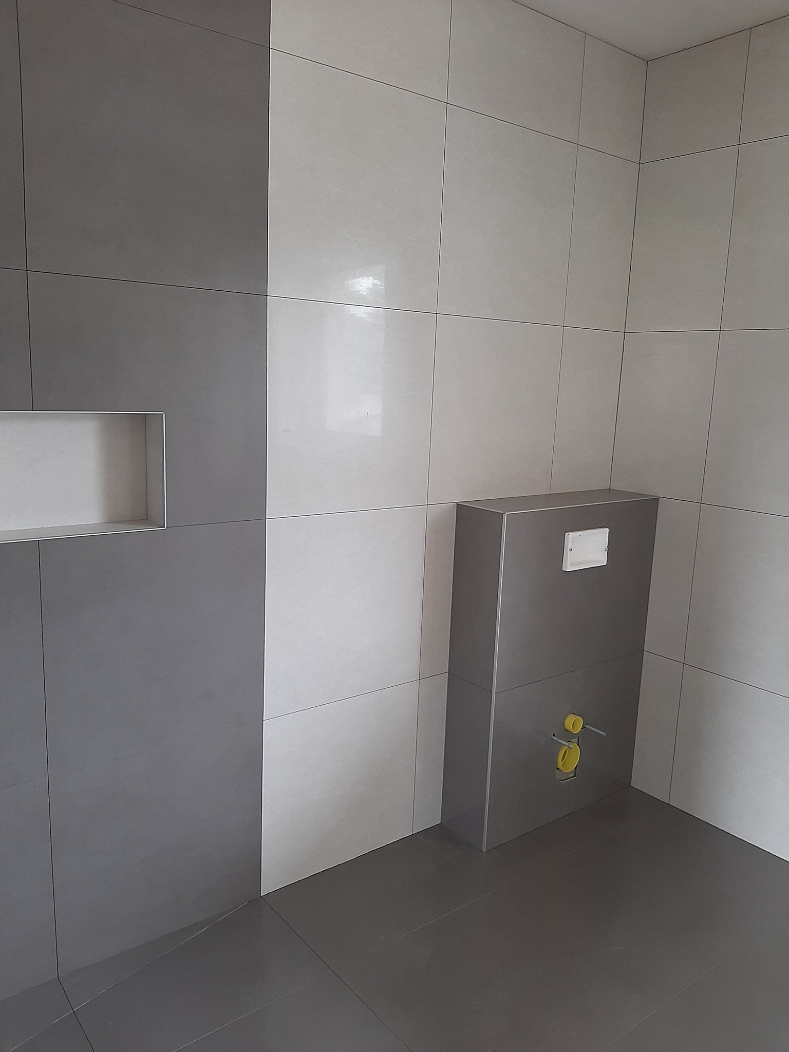 Nowoczesna łazienka z szarymi i białymi płytkami, zabudowa stelaża WC w kolorze szarym, wnęka w ścianie wykończona płytkami.