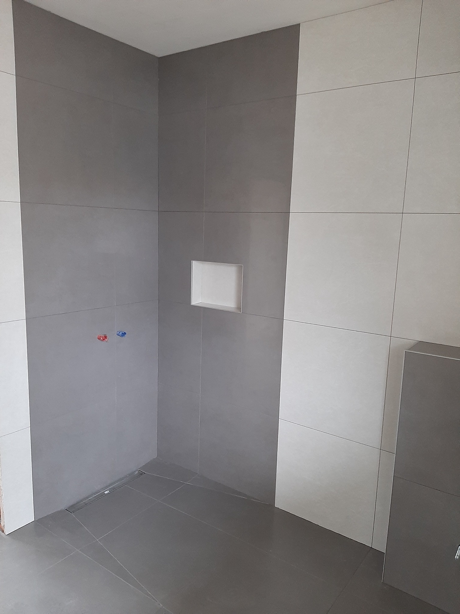 Świeżo wykafelkowana kabina prysznicowa z wnęką, wykończona w odcieniach szarości i bieli, widoczne przyłącza wodne i odpływ liniowy w podłodze.