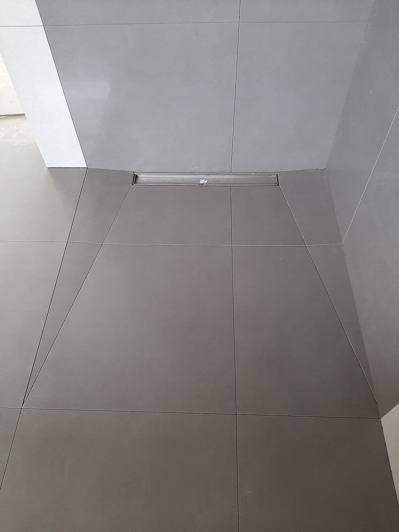 Szare, matowe płytki podłogowe ułożone ze spadkiem w kierunku liniowego odpływu prysznicowego ze stali nierdzewnej, widoczne fragmenty ściany i narożnika pomieszczenia.