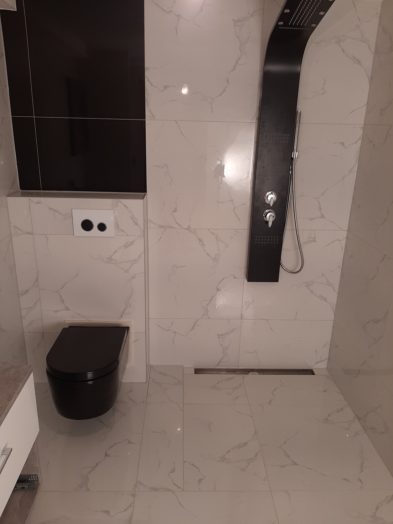 Nowoczesna łazienka z czarną armaturą: panel prysznicowy i wisząca toaleta, białe płytki imitujące marmur na ścianach i podłodze, widoczny odpływ liniowy.