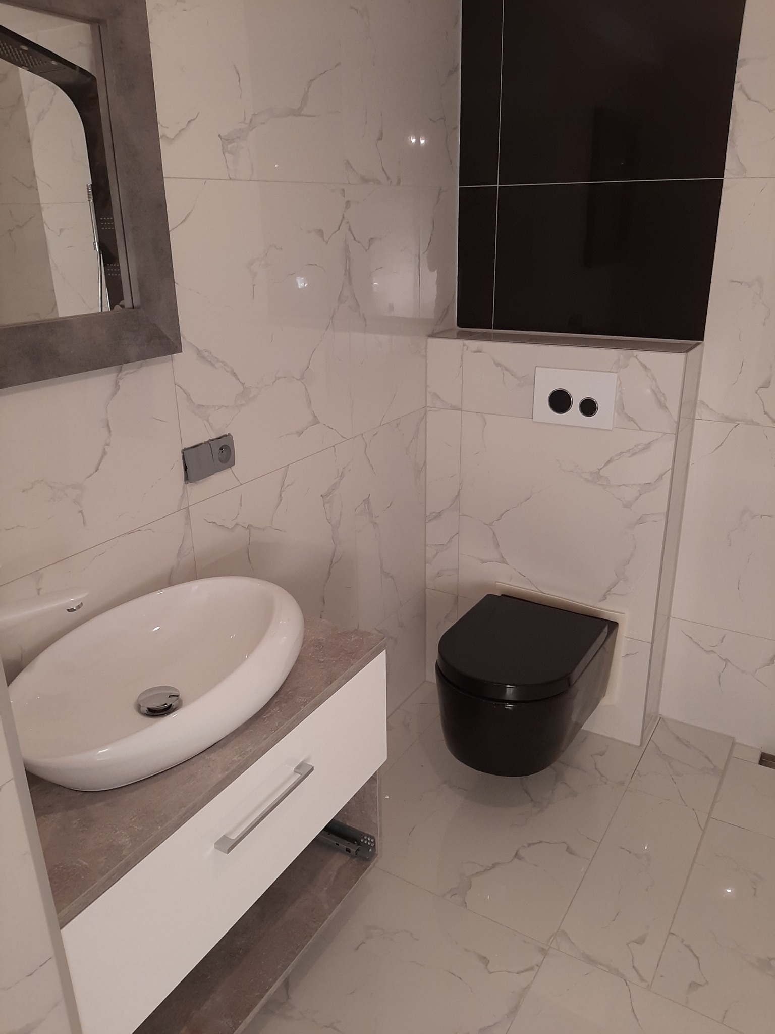 Nowoczesna łazienka z biało-szarymi marmurkowymi płytkami, wiszącą czarną toaletą i umywalką nablatową na białej szafce z szarym blatem, widoczne elementy armatury i lustro.