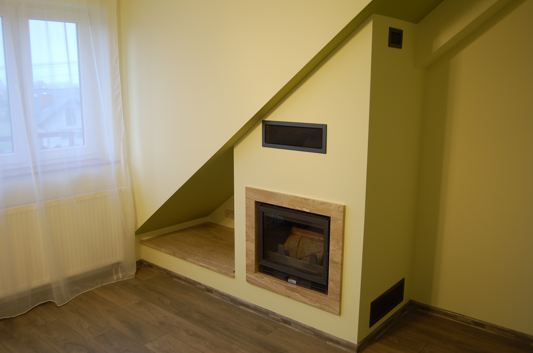 Narożnik pokoju z kominkiem obudowanym kamieniem, pod schodami, z drewnianą podłogą i grzejnikiem za firanką.