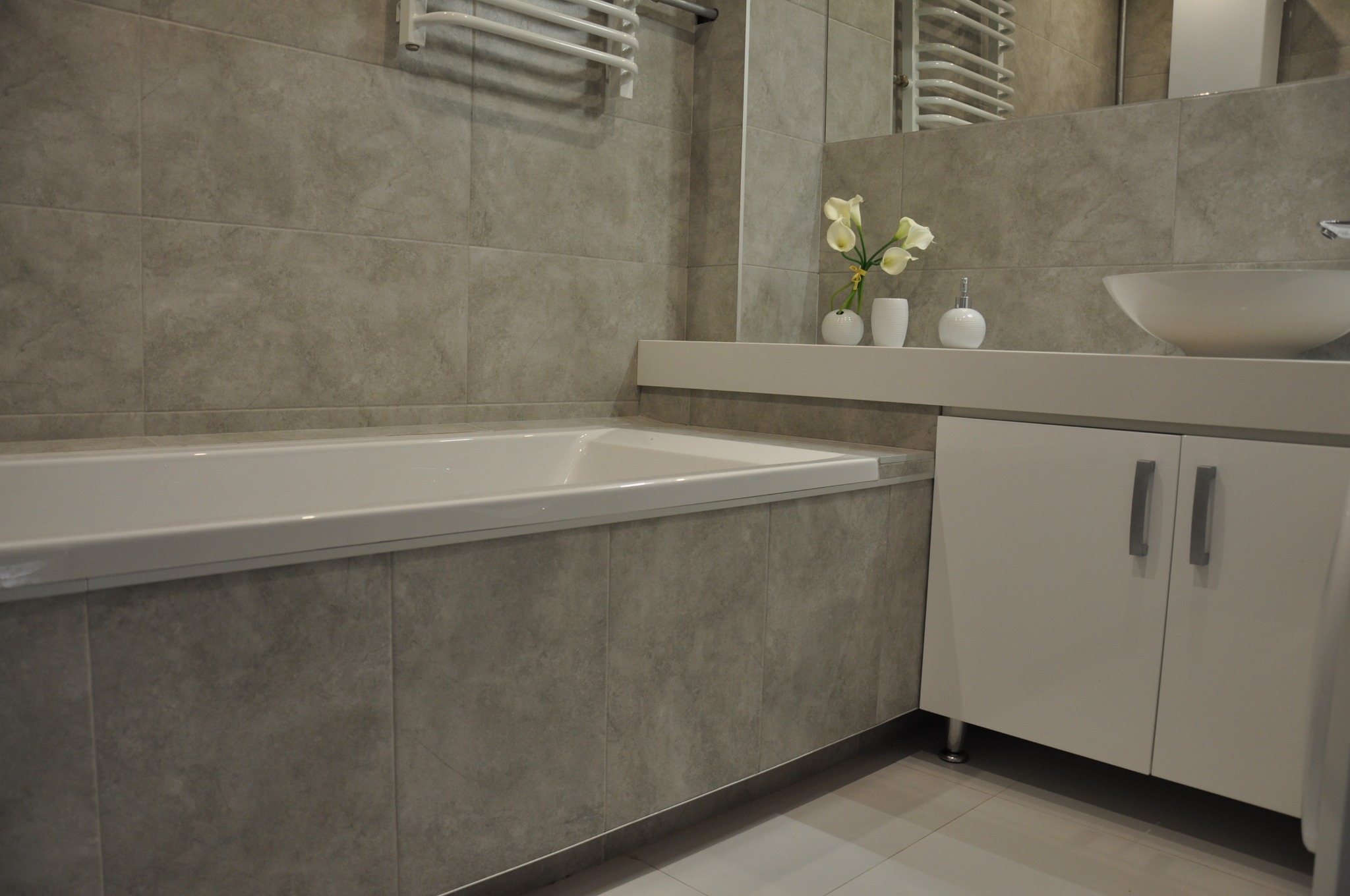 Wykończona łazienka z wanną obudowaną szarymi płytkami, białą szafką pod umywalką, kwiatami w wazonie i grzejnikiem drabinkowym.
