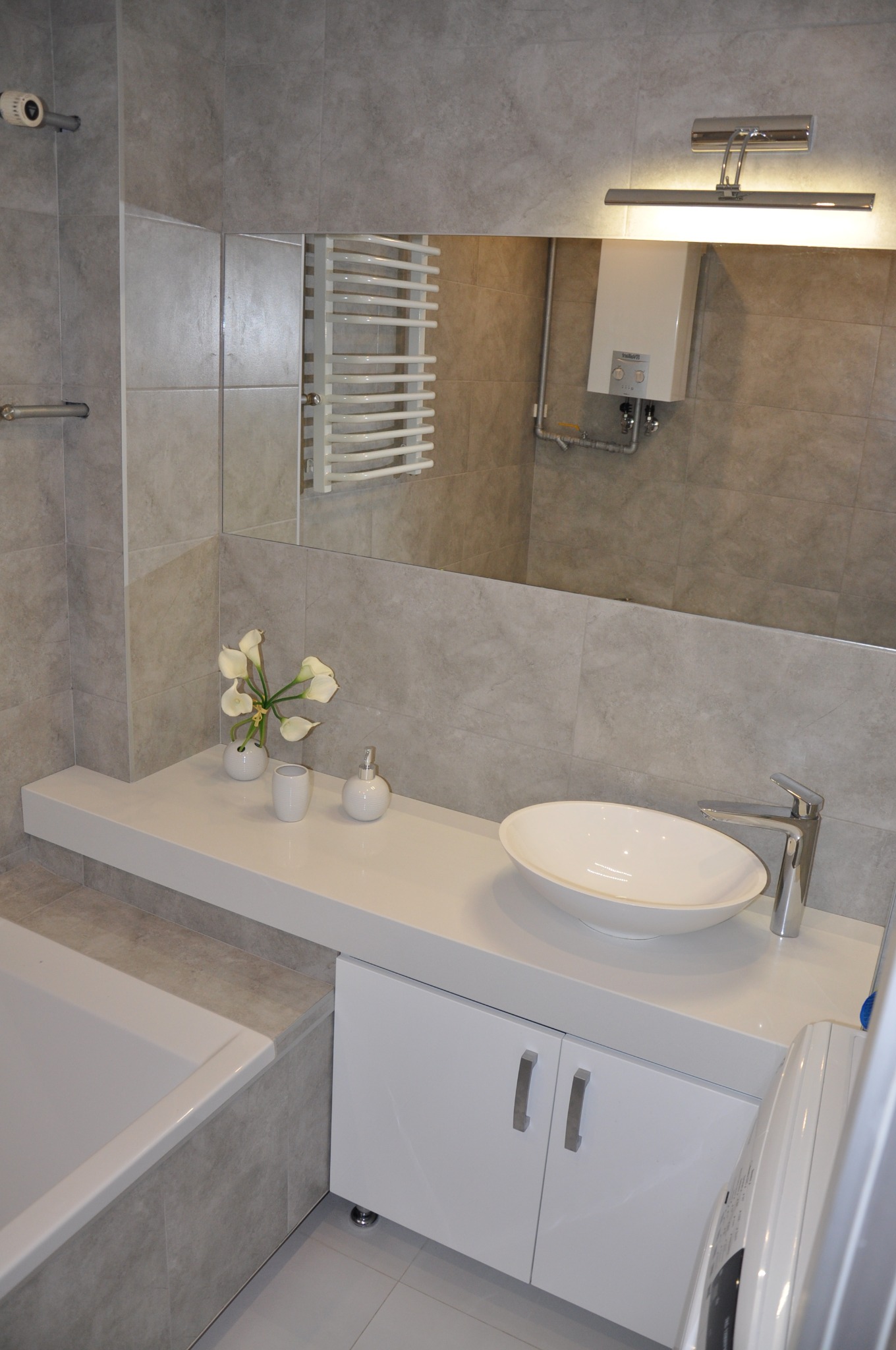 Jasna łazienka w odcieniach szarości z białą umywalką nablatową, baterią chromowaną, szafką pod umywalką i dekoracjami kwiatowymi, widoczne płytki ceramiczne na ścianach i podłodze oraz grzejnik...