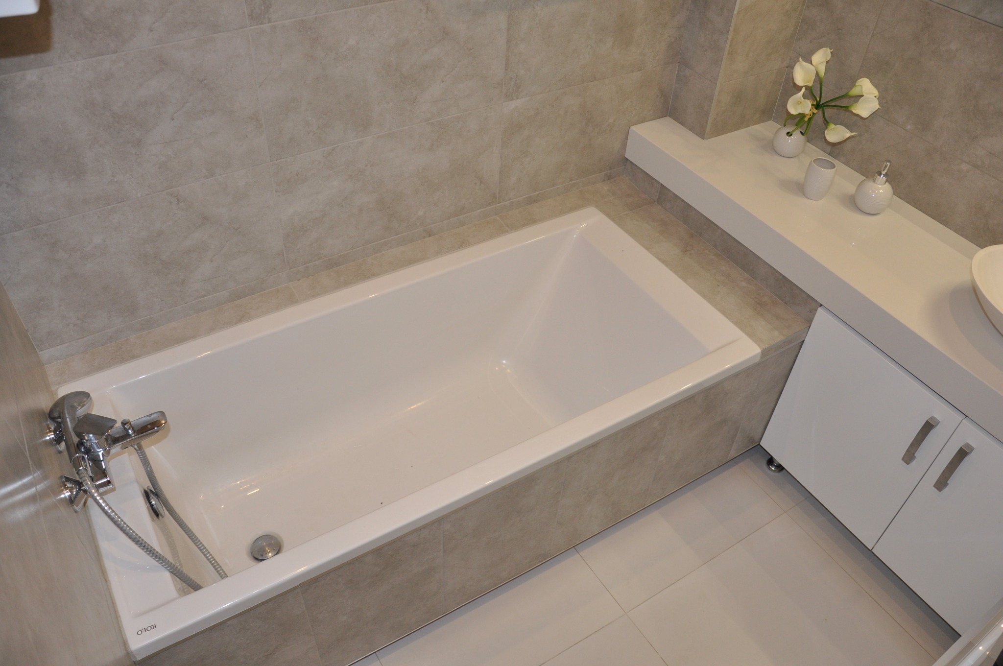 Biała wanna KOLO z chromowaną baterią, obudowana płytkami w odcieniach beżu, umywalka nablatowa, białe szafki pod umywalką, beżowe płytki na ścianach i podłodze, dekoracyjne białe kwiaty.