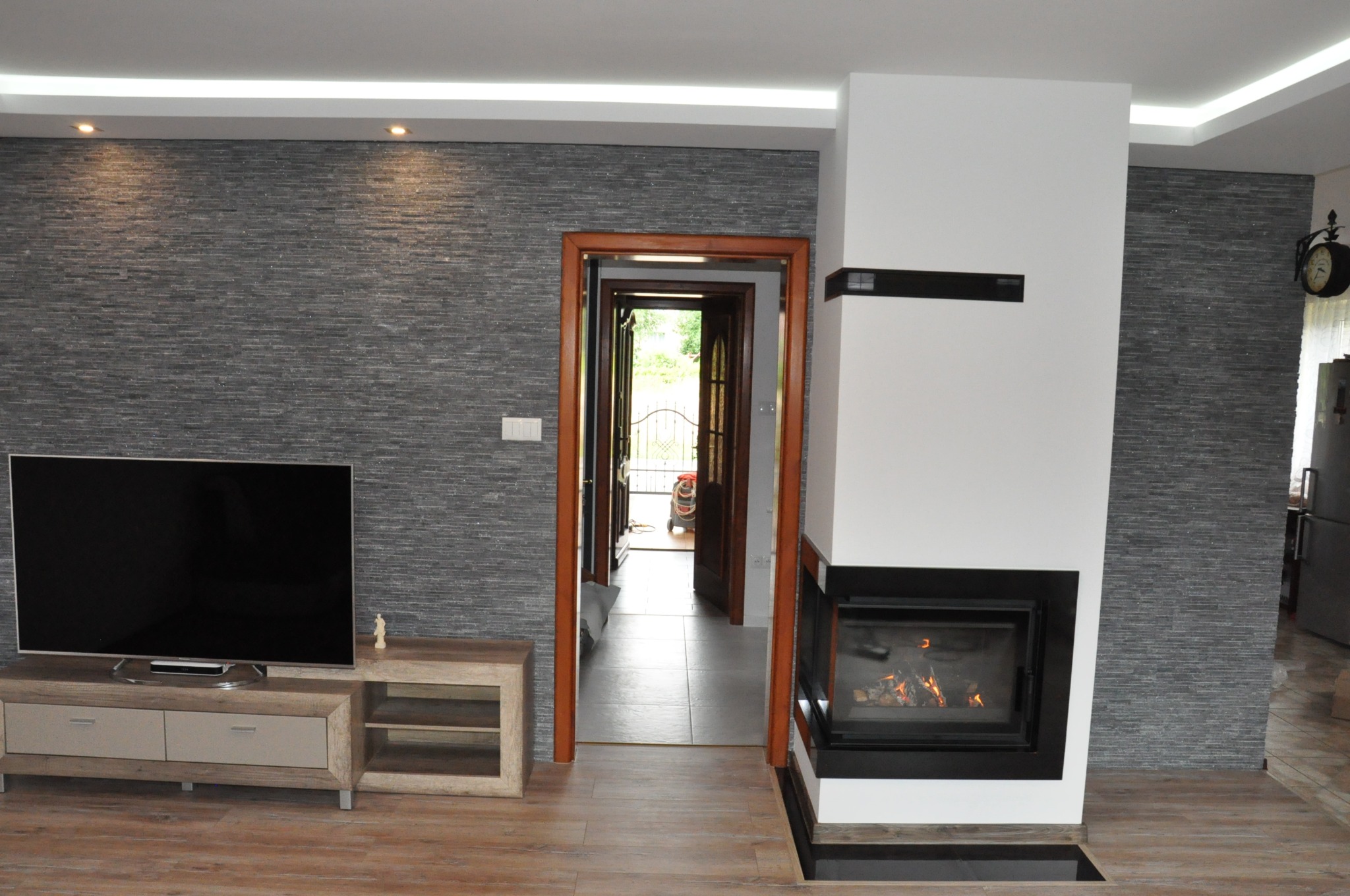 Salon z kominkiem obudowanym w białą płytę, ściana wykończona szarą okładziną imitującą kamień, drewniana podłoga i szafka RTV z telewizorem, widok na otwarte drzwi do dalszej części domu.