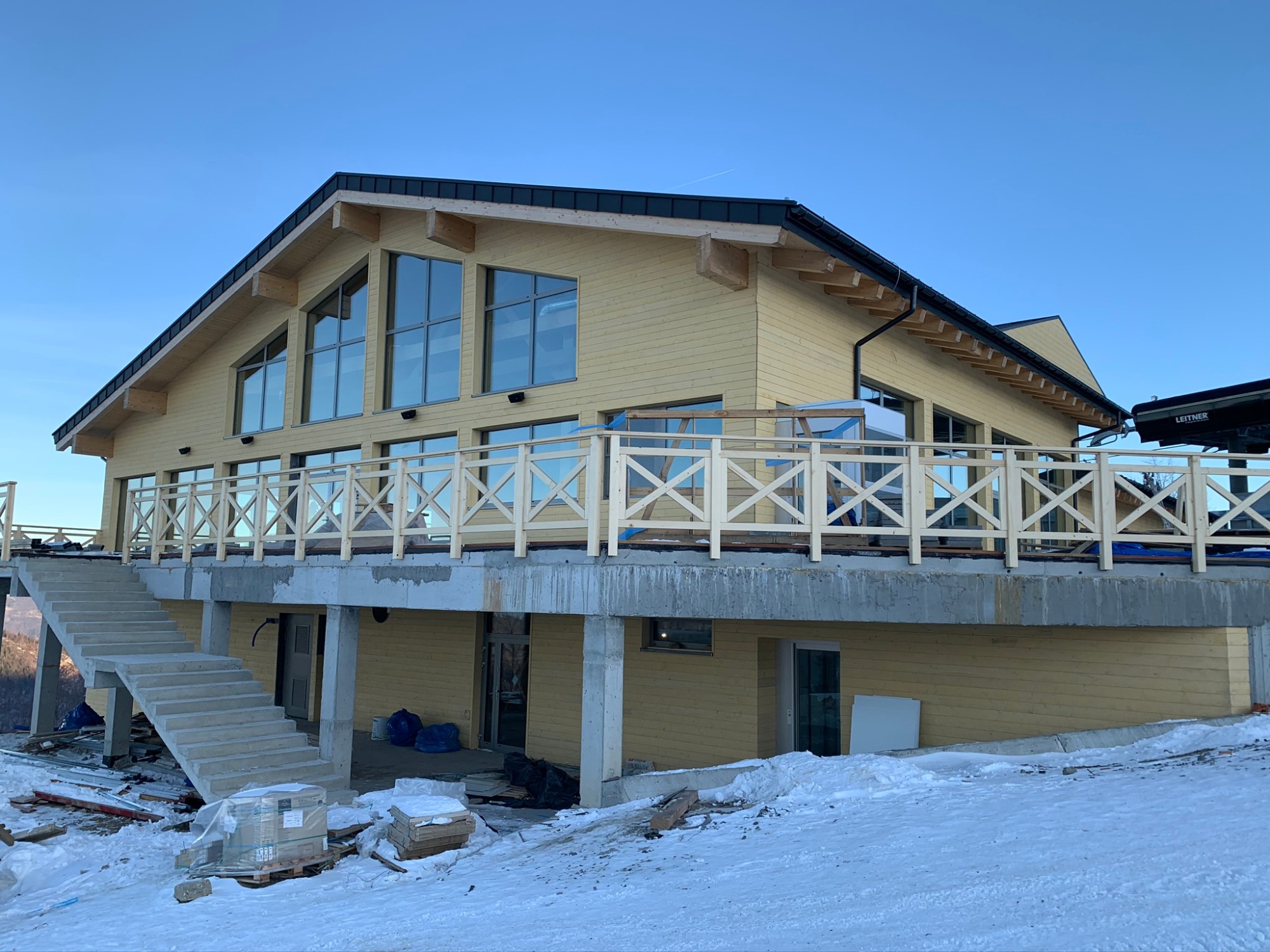 Żółty dom z drewnianą elewacją i dużymi oknami, z betonowymi schodami prowadzącymi na taras, w zimowym krajobrazie, częściowo w trakcie budowy.