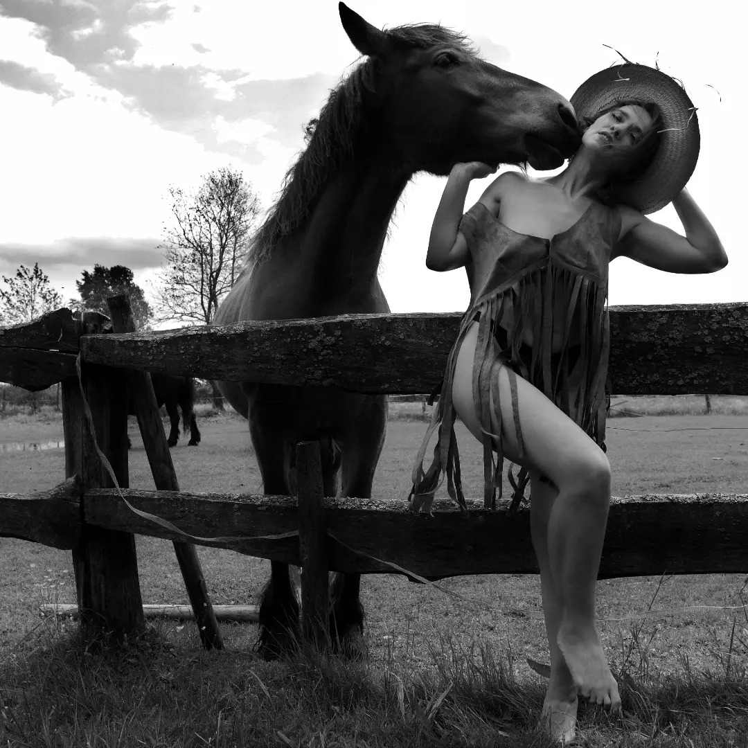 Czarno-białe zdjęcie kobiety w stroju z frędzlami i słomkowym kapeluszu, opierającej się o drewniany płot, z koniem dotykającym jej twarzy, w tle łąka i drzewa pod pochmurnym niebem.