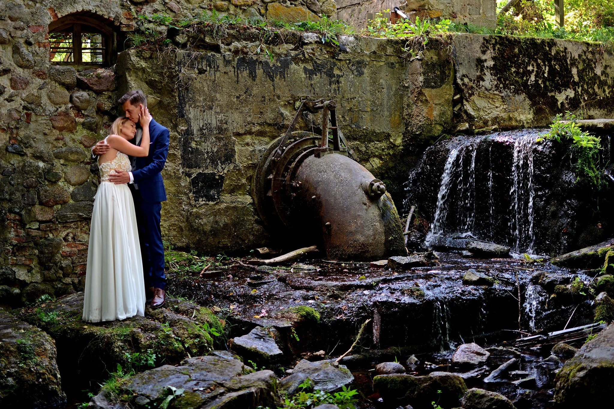 Para młoda w objęciach na tle starego, kamiennego młyna z wodospadem i zardzewiałą turbiną; panna młoda w długiej białej sukni z koronkową górą, pan młody w granatowym garniturze.