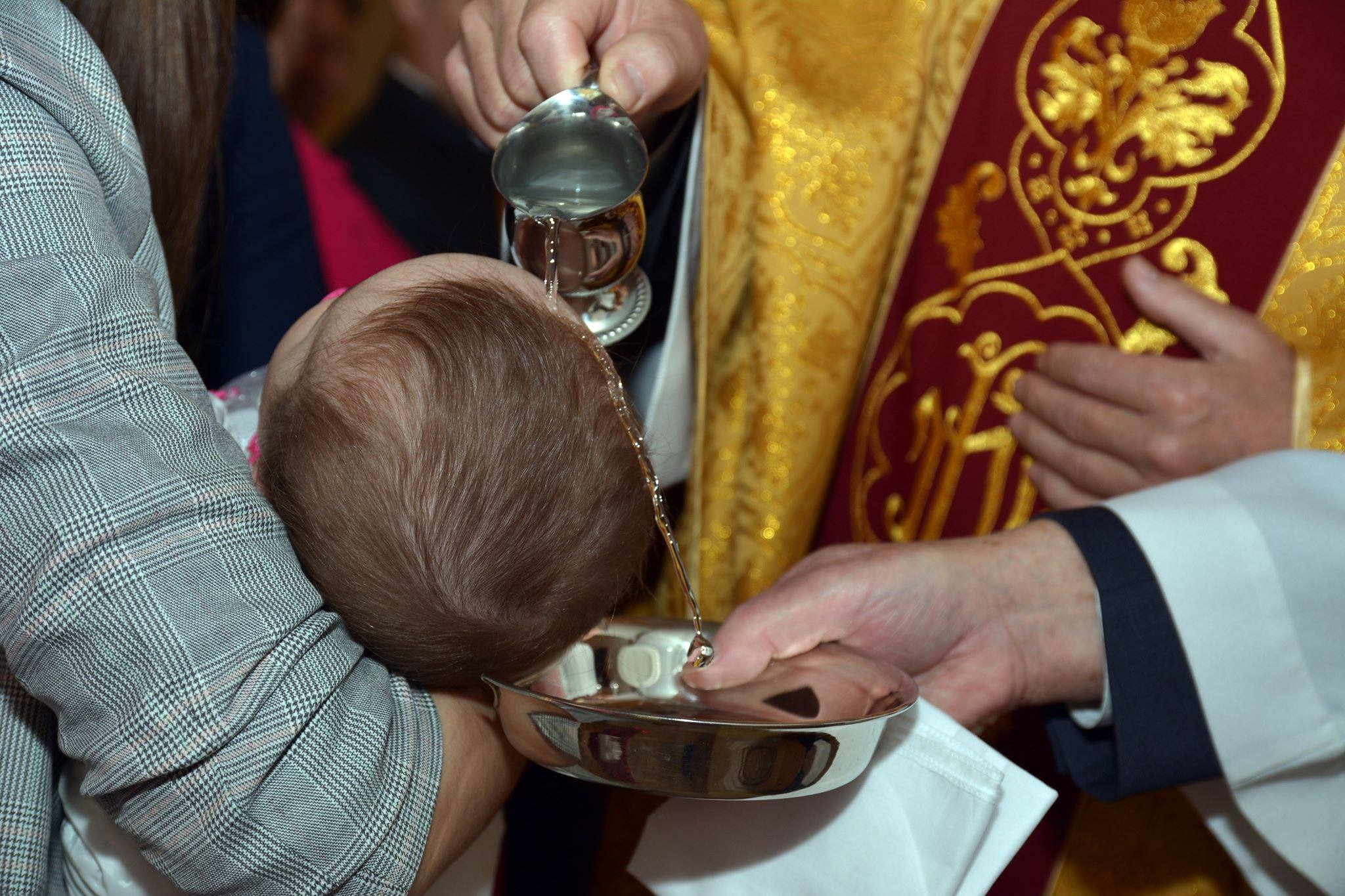 Chrzest dziecka: woda z naczynia polewana na główkę niemowlęcia trzymanego przez osobę w marynarce w kratę, podczas ceremonii w Suwałkach.