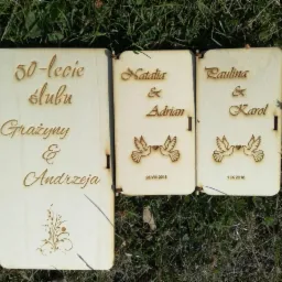 Drewniane, laserowo grawerowane pamiątki ślubne z imionami i datami, ułożone na trawie. Większa tabliczka upamiętnia 50-lecie ślubu.