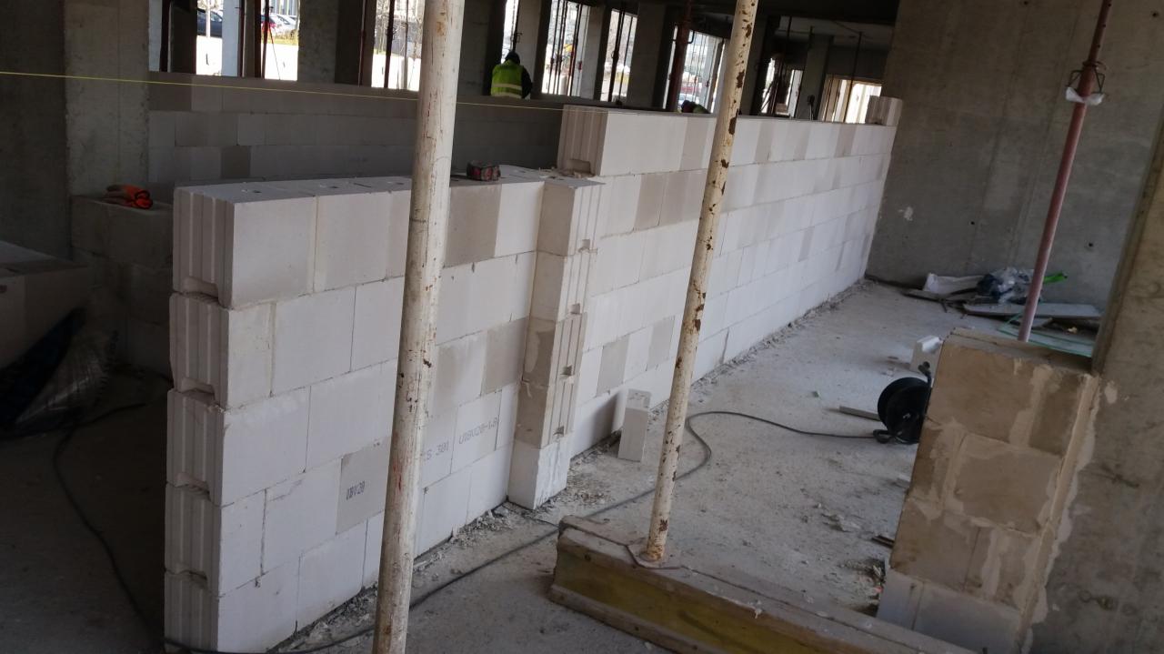 Wznoszenie ściany działowej z białych bloczków betonu komórkowego na budowie, widoczne stalowe podpory, okna w tle i częściowo ułożona instalacja elektryczna.