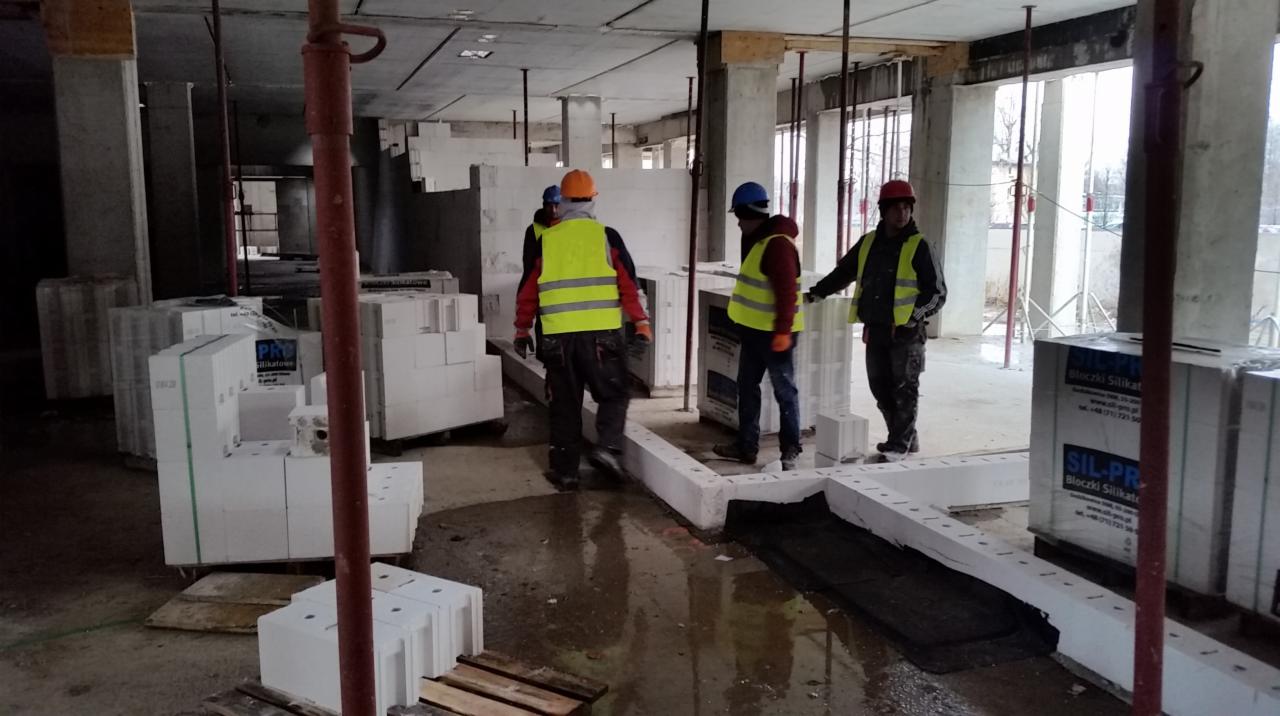 Wnętrze budynku w trakcie budowy z widocznymi pracownikami w kamizelkach odblaskowych, ułożonymi bloczkami silikatowymi oraz elementami konstrukcyjnymi, takimi jak podpory i filary.