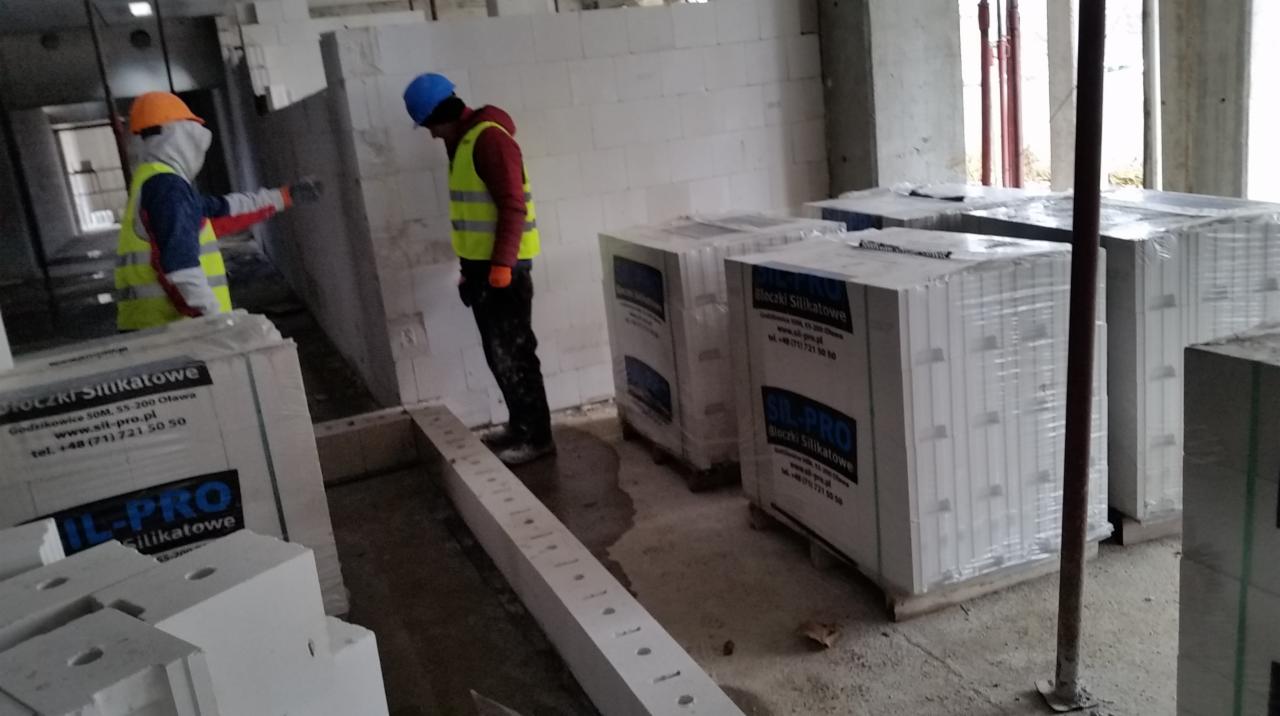 Budowa ścian z bloczków silikatowych; dwóch pracowników w kaskach i kamizelkach odblaskowych kontroluje jakość muru, obok palety z zapakowanymi bloczkami SIL-PRO.