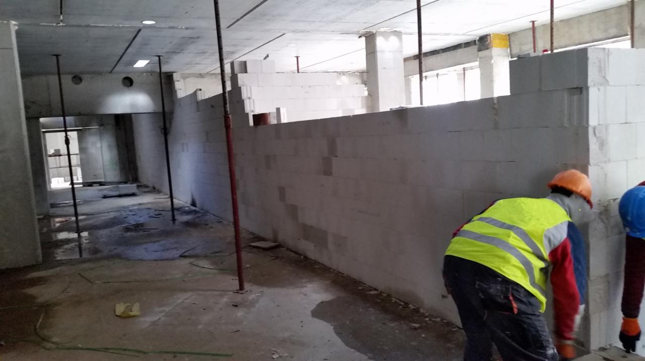 Wznoszenie ściany działowej z bloczków betonu komórkowego w surowym stanie, widoczni dwaj pracownicy w kaskach i kamizelkach odblaskowych, podpierające konstrukcje stalowe.