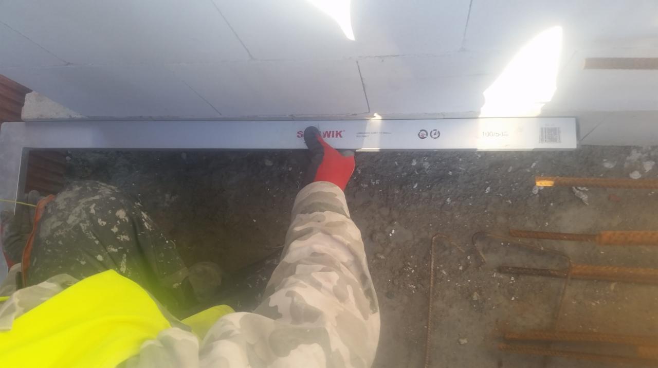 Pracownik budowlany w odblaskowej kamizelce i rękawicy mierzy poziom wylewki betonowej przy użyciu poziomicy 'S Wik' wewnątrz fundamentów pod ściany z betonu komórkowego.