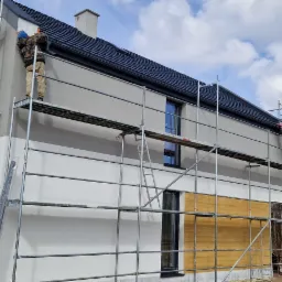 Dwukondygnacyjny dom w trakcie tynkowania elewacji, widoczne rusztowania, pracownicy w kombinezonach, częściowo otynkowana ściana w dw&oacute;ch odcieniach szarości i bieli, okna zabezpieczone folią,...