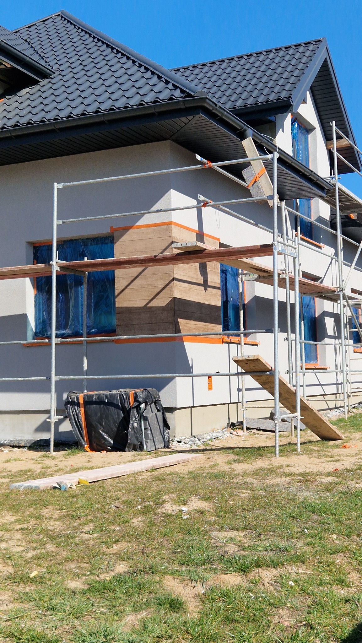 Nowy dom w trakcie wykańczania elewacji, widoczne rusztowania, okna zabezpieczone folią, częściowo nałożony dekoracyjny panel elewacyjny imitujący drewno, na ziemi czarna folia budowlana.