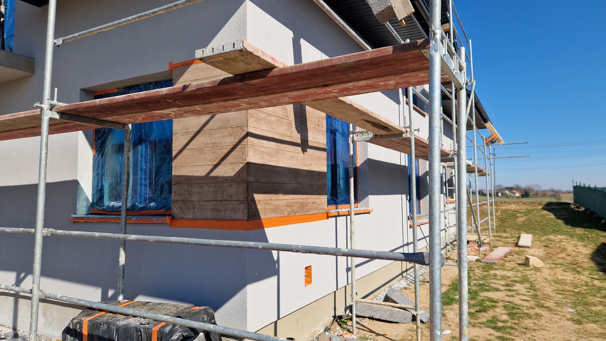 Fragment elewacji domu w trakcie prac wykończeniowych, z widocznymi rusztowaniami, oknami zabezpieczonymi folią i częściowo wykończoną ścianą imitującą drewno.