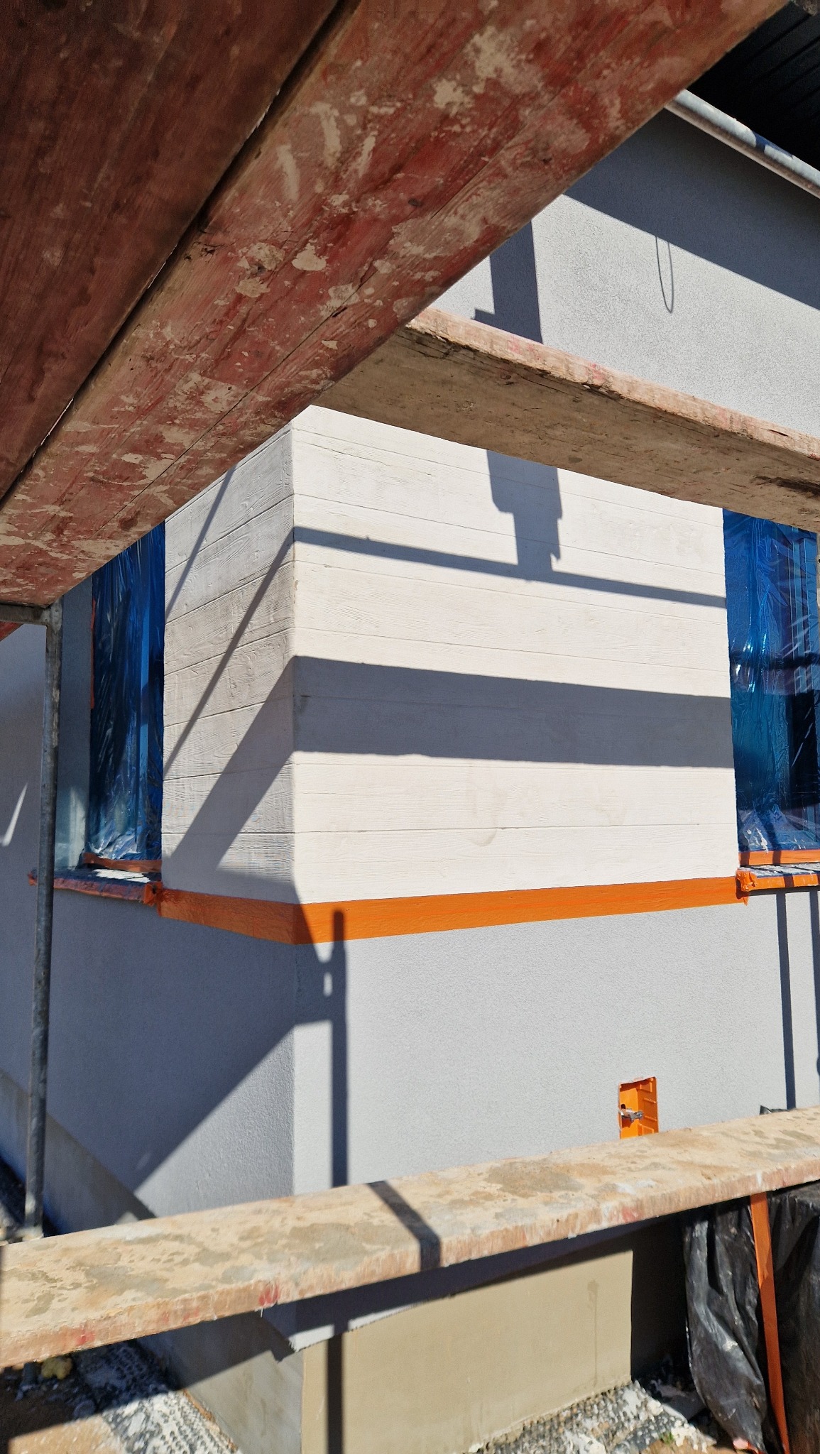 Fragment elewacji budynku w trakcie prac, z widocznym rusztowaniem, zabezpieczonym oknem folią i pomalowanym na pomarańczowo cokołem.