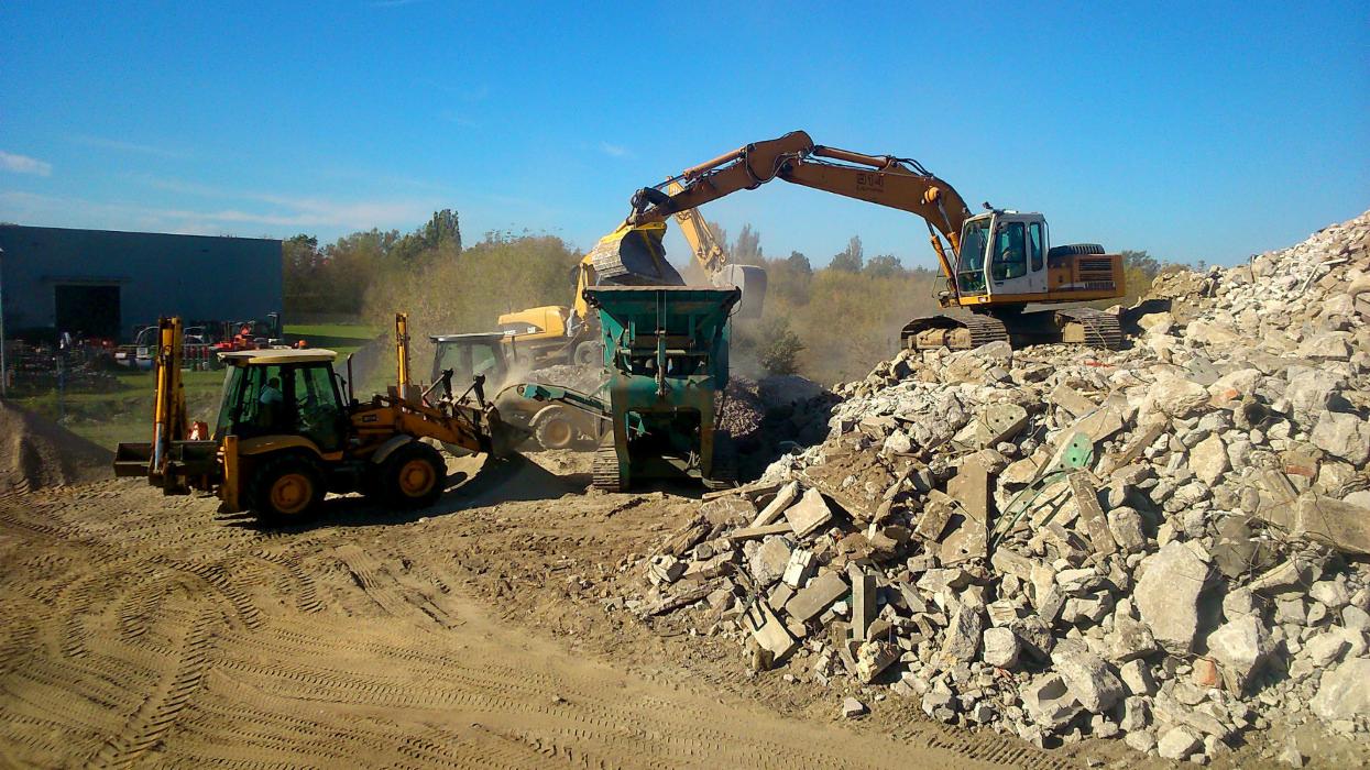 Żółta koparka z łyżką umieszcza gruz w zielonym kruszarce na gąsienicach, w tle widoczny budynek i inne maszyny budowlane, ziemia pokryta śladami opon.