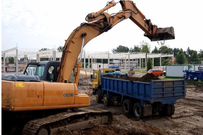 Pomarańczowa koparka CX210 nabiera ziemię i wsypuje ją do niebieskiej ciężarówki na placu budowy, w tle widoczny szkielet budowanego budynku.