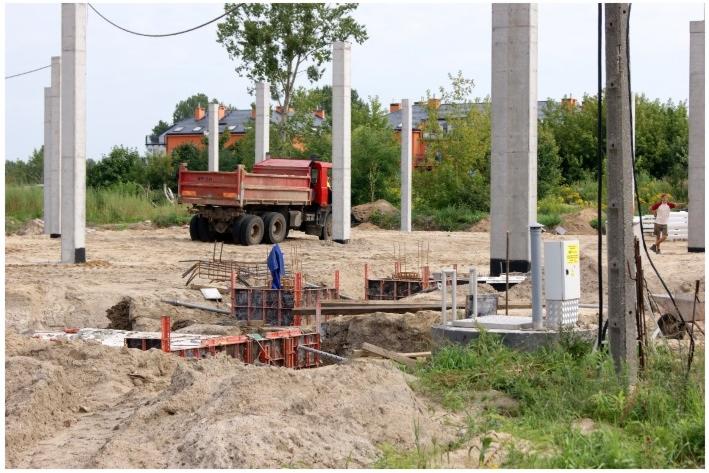 Budowa fundamentów hali: betonowe słupy, szalunki zbrojeniowe, wykop z widoczną instalacją, ciężarówka w tle.