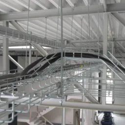 Instalacja kabli elektrycznych w korytach siatkowych pod sufitem w hali przemysłowej, widoczne rury i stalowa konstrukcja nośna.