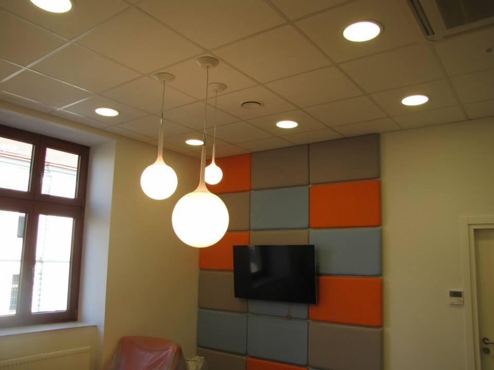 Wnętrze biurowe z trzema kulistymi lampami wiszącymi o różnej wielkości, podwieszanym sufitem z wbudowanymi oprawami, oknem z ciemną ramą i ścianą z panelami akustycznymi w odcieniach szarości...