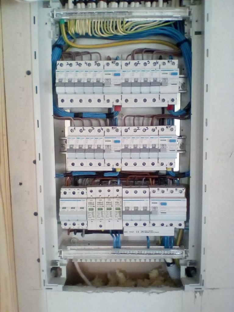 Rozdzielnica elektryczna z zainstalowanymi zabezpieczeniami różnicowoprądowymi i nadprądowymi, widoczne przewody w kolorach niebieskim, żółto-zielonym i brązowym, częściowo osłonięta wełną mineralną.