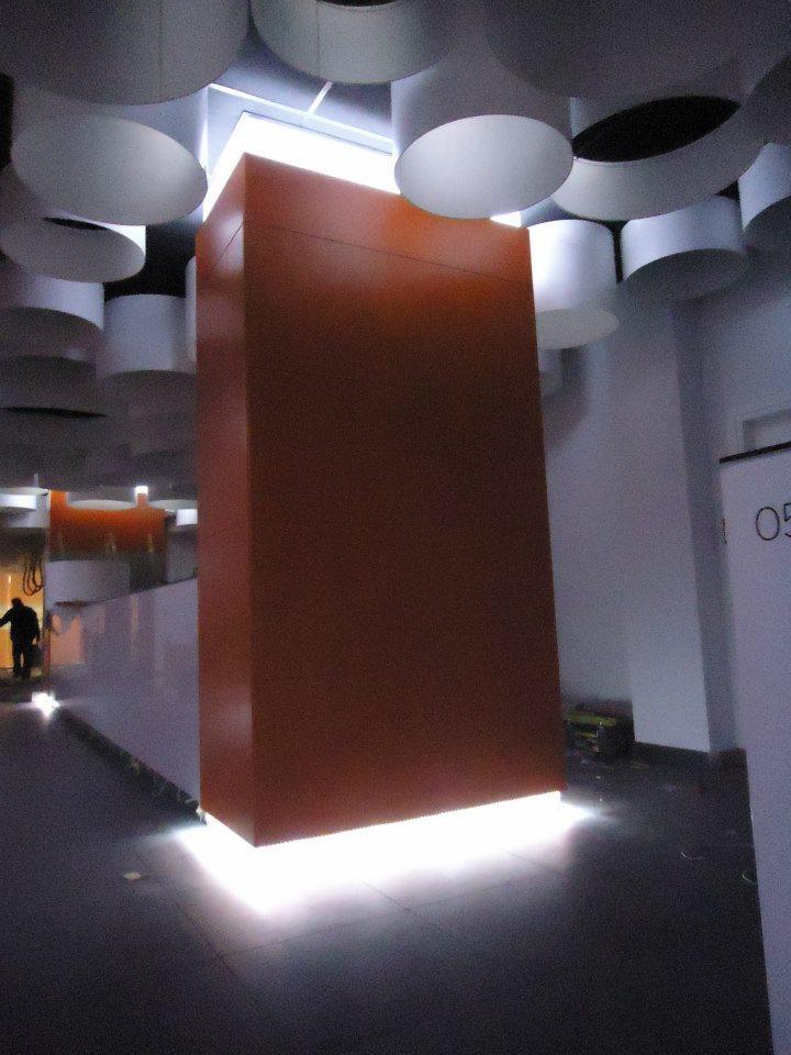 Wysoka, brązowa kolumna oświetlona od dołu białym światłem LED, otoczona cylindrycznymi lampami sufitowymi w nowoczesnym wnętrzu. W tle widoczna sylwetka osoby.