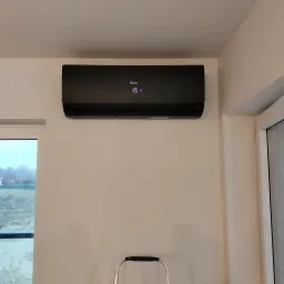 Czarny klimatyzator Haier zamontowany na jasnej ścianie w nowym budynku, widoczny fragment drabiny i okno z widokiem na plac budowy.