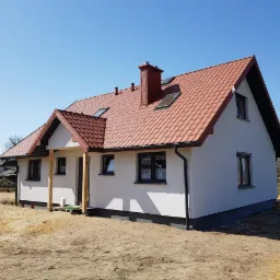 Nowy dom jednorodzinny z czerwoną dachówką i świeżo otynkowaną, białą elewacją, widoczne przygotowania do wykończenia werandy.