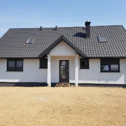 Nowoczesny dom jednorodzinny z białą elewacją, grafitowym dachem z dachówki, czarnymi oknami z roletami zewnętrznymi i charakterystycznymi elementami wykończeniowymi, takimi jak czarny komin...