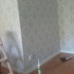 Fragment pokoju po położeniu jasnej tapety w delikatny wzór, widoczna drabina malarska, tuba z klejem i narzędzia na drewnianej podłodze.