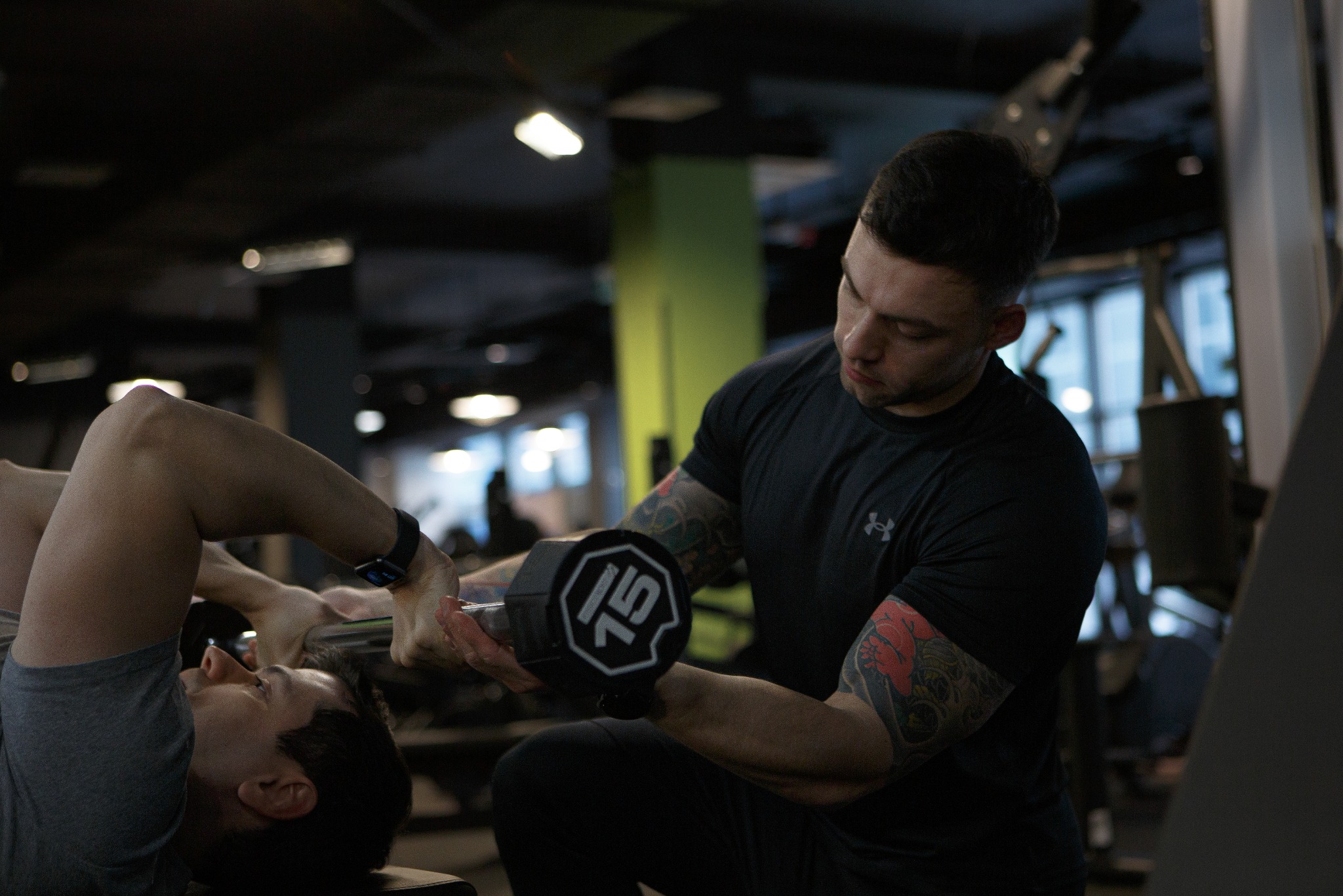 Mężczyzna z tatuażami na ramionach asekuruje ćwiczącego mężczyznę leżącego na ławce, który podnosi sztangielkę o wadze 15 kg w siłowni.