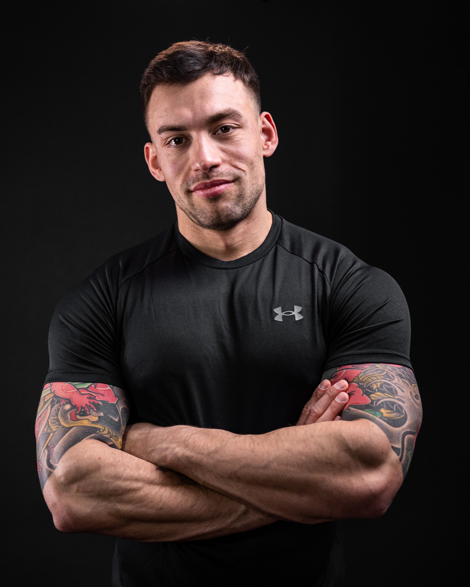Portret mężczyzny w czarnej koszulce sportowej z tatuażami na ramionach, skrzyżowane ręce na tle ciemnego studia.