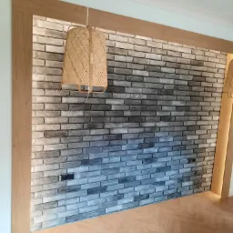 Ściana z szarej cegły dekoracyjnej w drewnianej ramie, oświetlona wiszącą lampą z abażurem z naturalnego materiału, nad parkietem ułożonym w jodełkę.