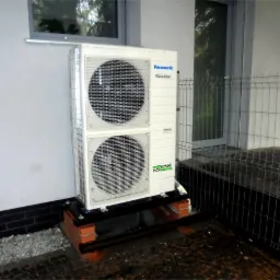 Zewnętrzna jednostka pompy ciepła Panasonic T-CAP zamontowana na ceglanej podstawie przy ścianie budynku z widocznym oknem i metalowym ogrodzeniem.