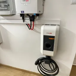 Biała stacja ładowania z napisem 'Insert' na ścianie, z podłączonym kablem. Obok widoczny inwerter z przewodami.