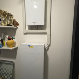 Solcity - Fotowoltaika i Systemy Grzewcze - Dwa białe moduły ATMOCE zamontowane na ścianie w garażu, połączone listwami. Górny moduł z okrągłym wskaźnikiem, dolny z niebieską diodą. Obok półki z trofeami i środkami czystości.