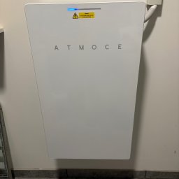 Solcity - Fotowoltaika i Systemy Grzewcze - Biały, trapezoidalny magazyn energii ATMOCE na ścianie, z niebieską diodą i żółtą naklejką ostrzegawczą. Widoczny fragment instalacji elektrycznej i podłogi.